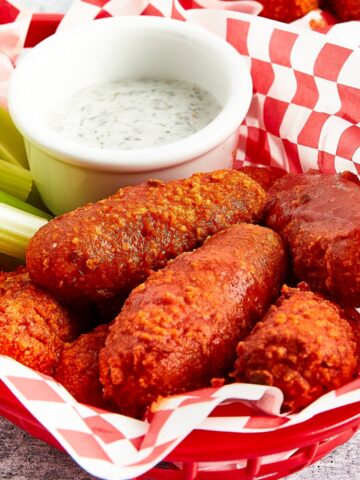 Bright orange gluten free wings sit in a red plastic serving basket lined with red and white checkered paper. Celery sticks and a small white bowl of celery ranch are in the basket with the wings.