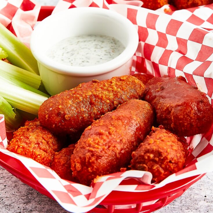 Bright orange gluten free wings sit in a red plastic serving basket lined with red and white checkered paper. Celery sticks and a small white bowl of celery ranch are in the basket with the wings.