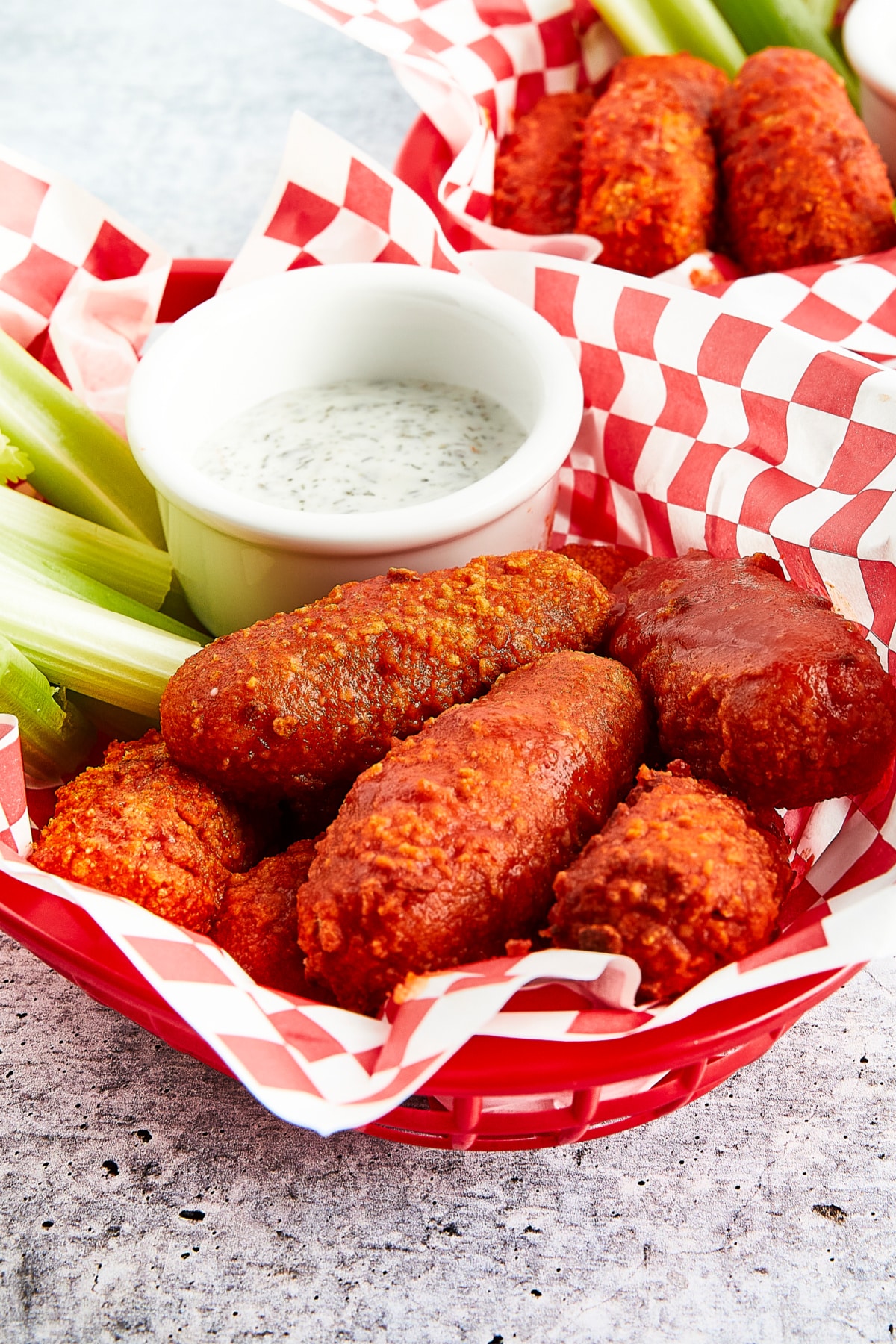 Bright orange vegan buffalo wings sit in a red plastic serving basket lined with red and white checkered paper. Celery sticks and a small white bowl of celery ranch are in the basket with the wings.