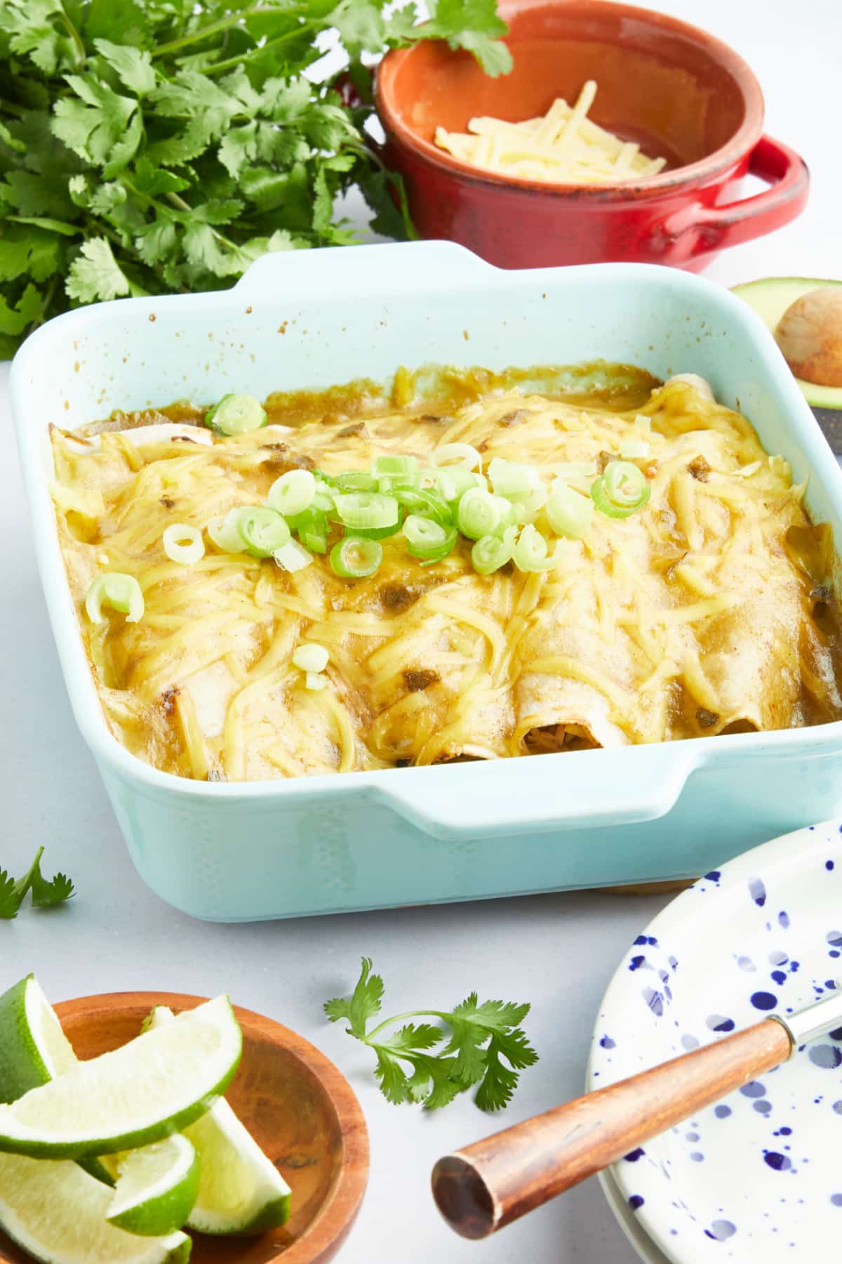 A baking dish of enchiladas sit on a white tabletop next to a stack of serving plates, bowls of lime wedges and shredded cheese, and a bunch of parsley.