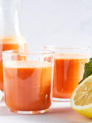 Side view of two glasses of bright orange juice and a larger bottle of juice. Two lemon halves, a tree of fresh broccoli, and a garlic bulb sit next to the juice.