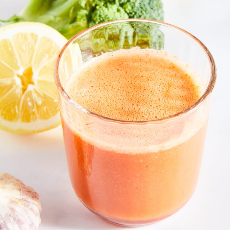 Close up of one glass of bright orange juice. Half of a lemon, a large broccoli tree, and a garlic bulb sit next to the juice.