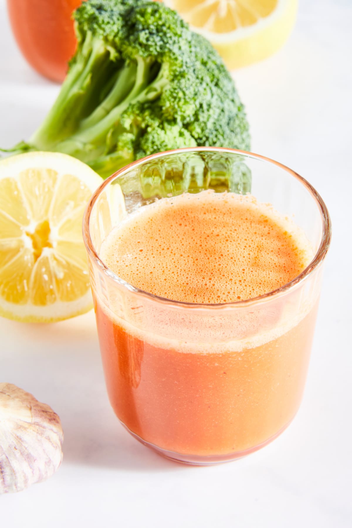 One glass of bright orange immune boosting juice sits on a bright white surface. Two lemon halves, a large broccoli tree, and a garlic bulb sit next to the juice.