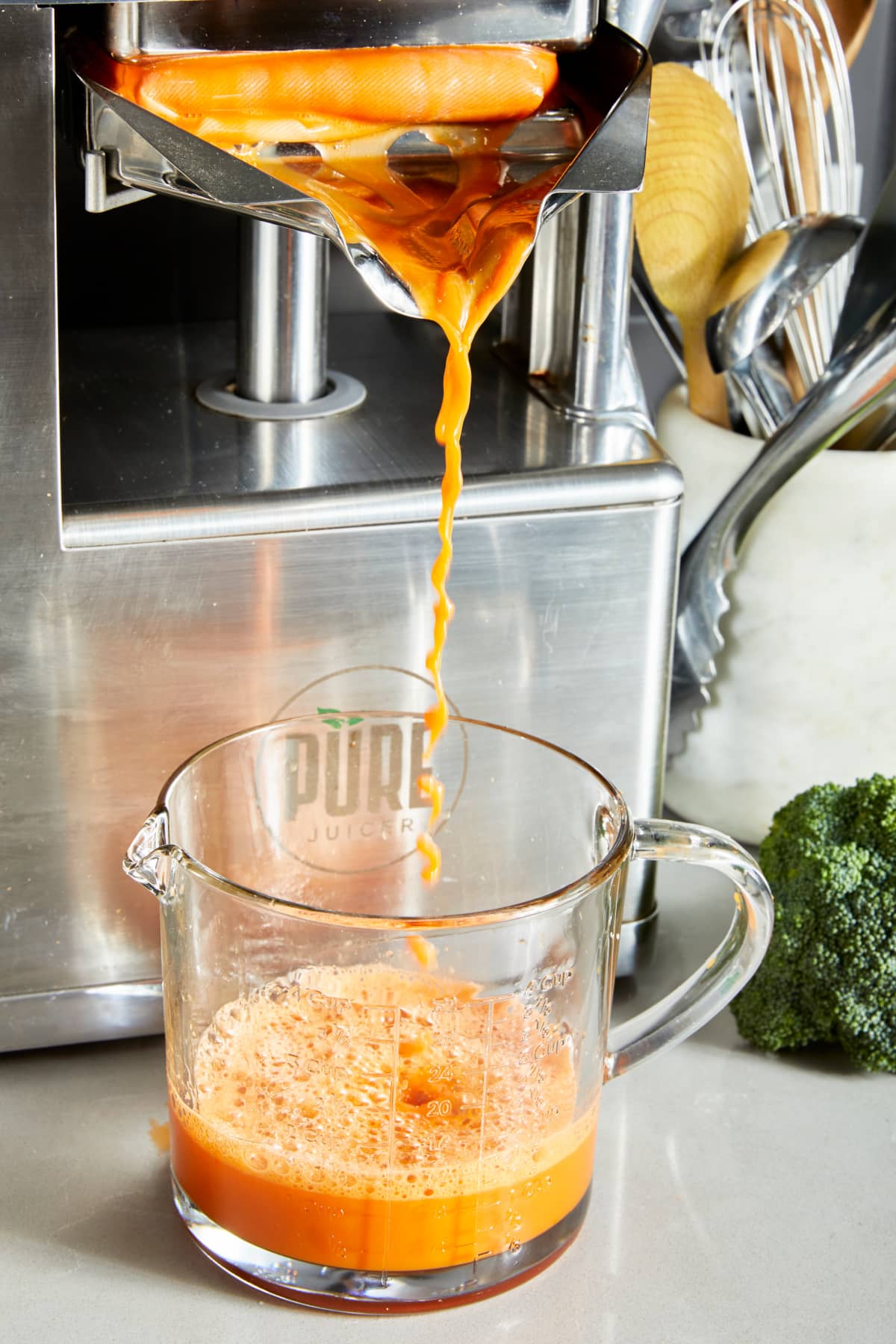 A brushed stainless steel juicer pours bright orange immune boosting juice out into a glass pitcher with a handle.
