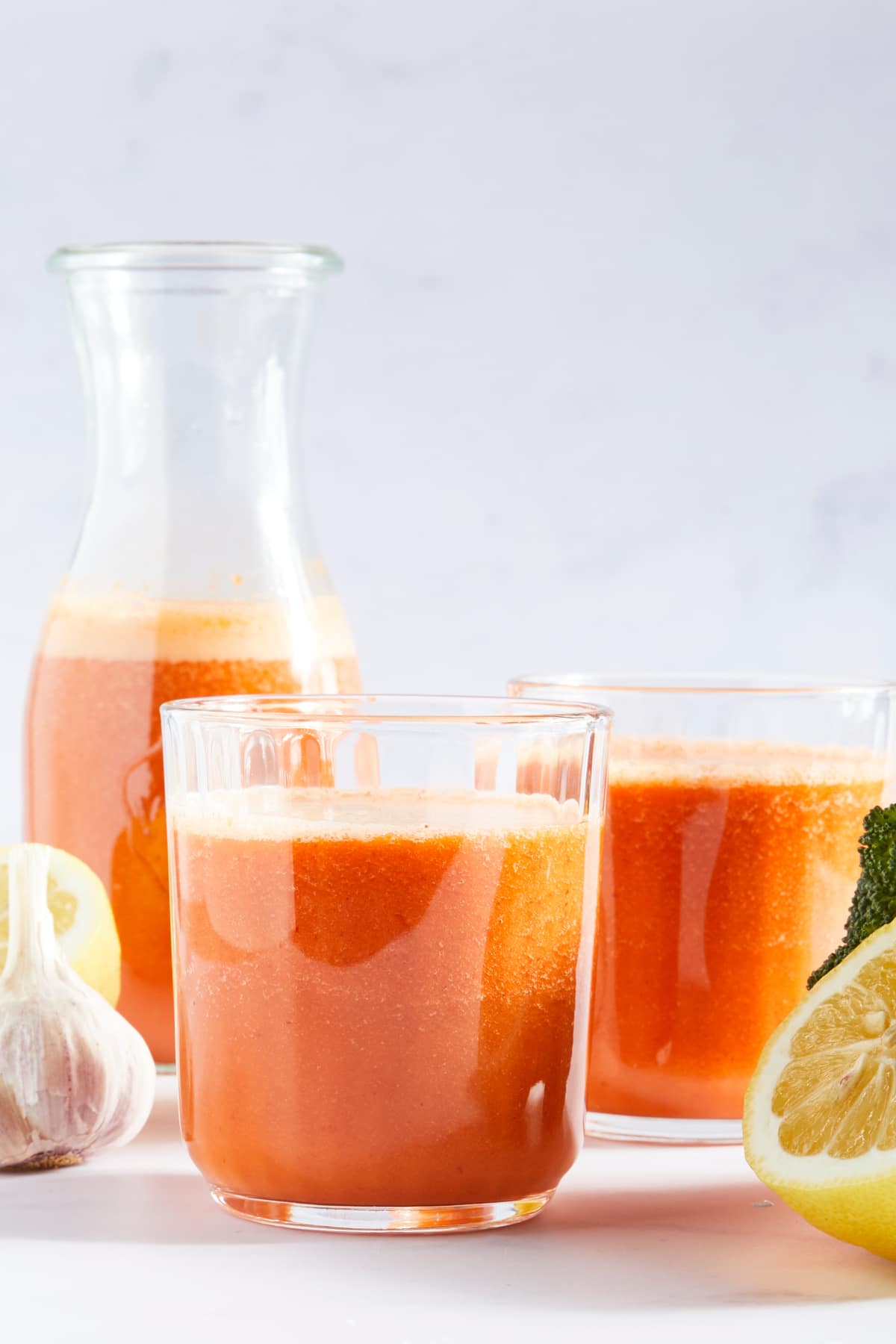 Side view of two glasses of bright orange juice and a larger bottle of juice. Two lemon halves and a garlic bulb sit next to the juice.
