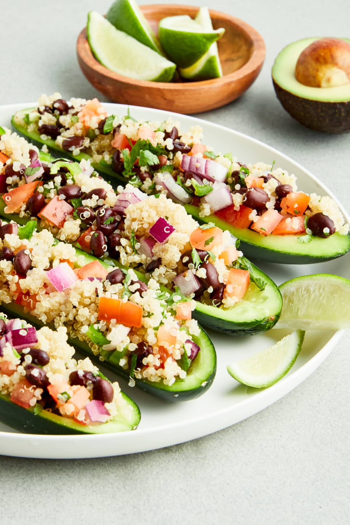 Four cucumber boats filled with a tex mex mixture of beans, quinoa, tomato and onion sit on a white plate. A small wooden bowl of lime wedges and a half avocado sit next to the plate.