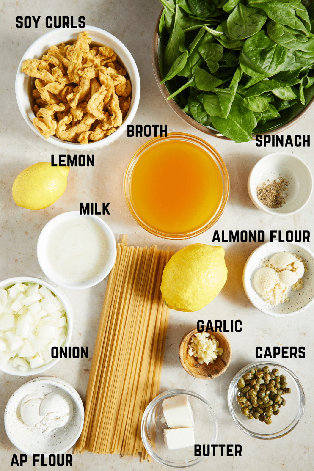 Overhead view of ingredients to make vegan chicken piccata: soy curls, spinach, lemons, broth, spices, almond flour and AP flour, milk, garlic, onion, capers, pasta noodles, and butter.