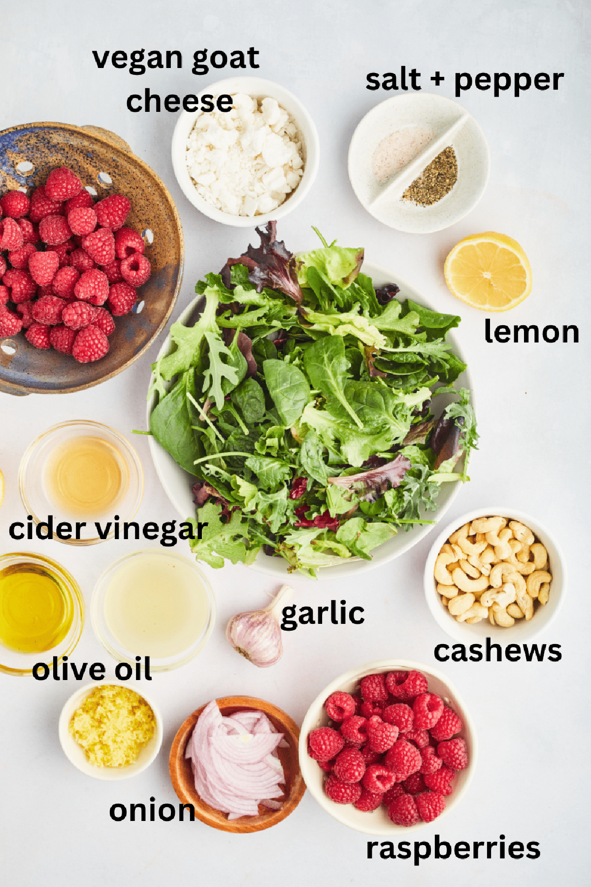 Overhead view of ingredients to make a roasted garlic raspberry salad: mixed greens, vegan goat cheese, fresh raspberries, salt and pepper, lemons, cashews, garlic, apple cider vinegar, olive oil, and red onion.