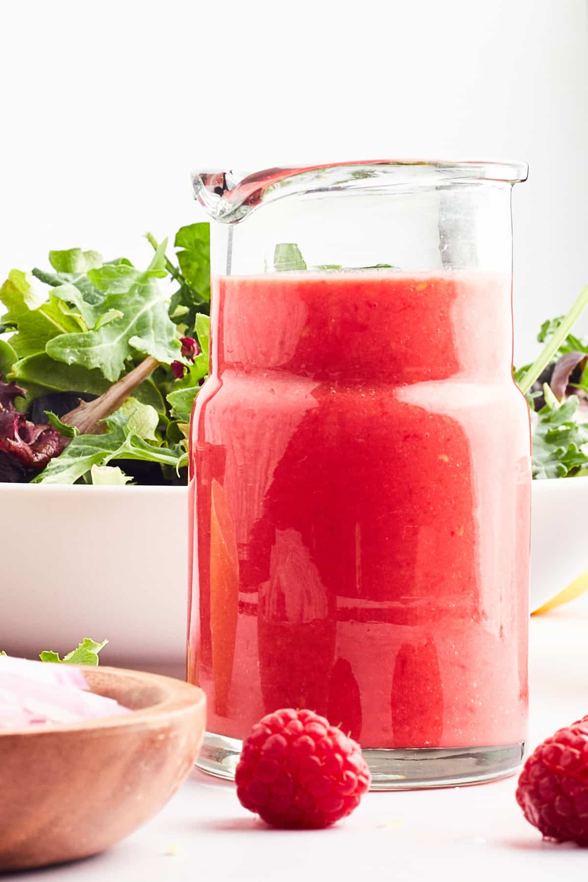 A small glass pitcher of bright red raspberry vinaigrette sits in front of a large bowl of salad greens.