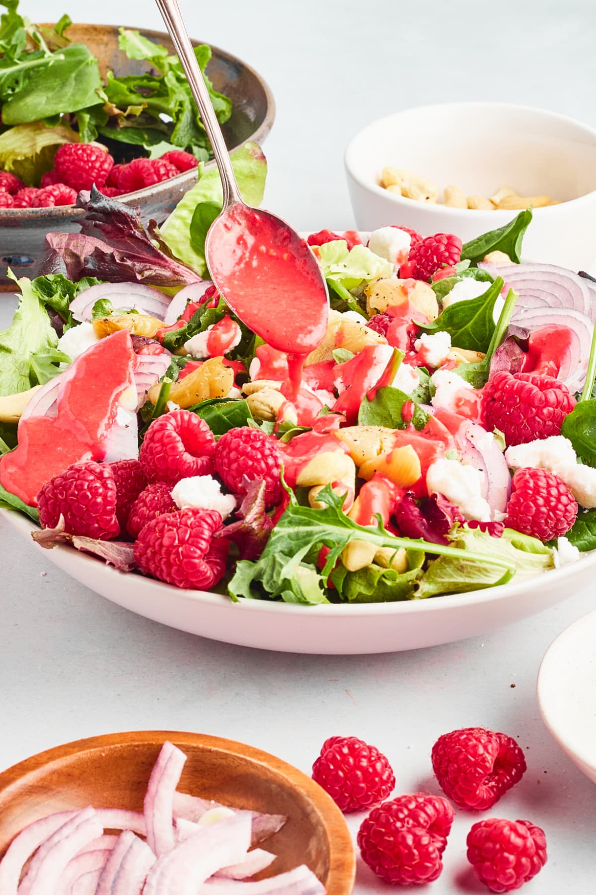 A mixed greens salad with roasted garlic and fresh raspberries. The salad is served in a large white bowl and sitting on a white surface, and a spoon is drizzling a bright red raspberry vinaigrette over the salad. A rustic blue colander holds more greens and berries and sits next to the salad. Small bowls hold sliced red onion, cashews, and vegan goat cheese.
