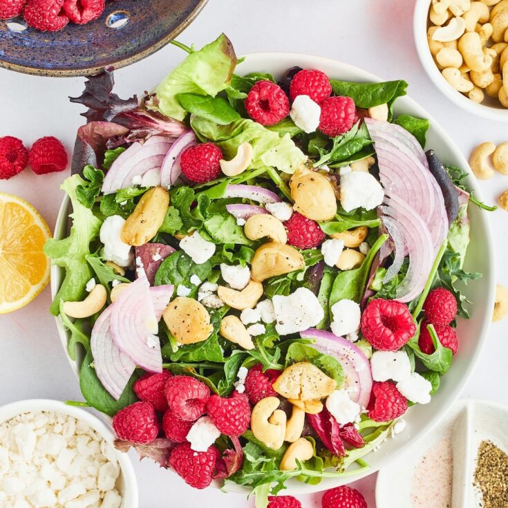 An overhead view of a mixed greens salad with roasted garlic and fresh raspberries. The salad is served in a large white bowl and sitting on a white surface. A rustic blue colander holds more greens and berries and sits next to the salad. Small bowls hold sliced red onion, cashews, and vegan goat cheese.