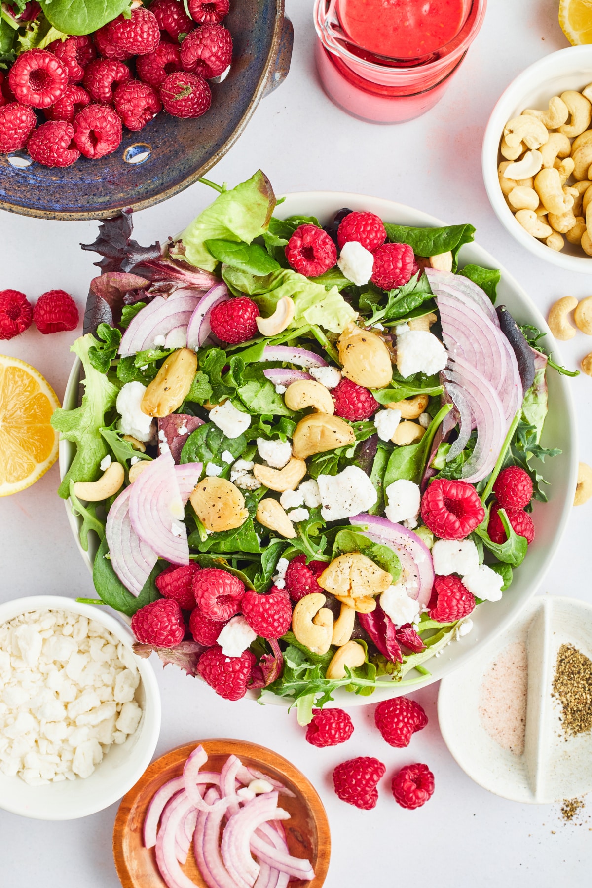 An overhead view of a mixed greens salad with roasted garlic and fresh raspberries. The salad is served in a large white bowl and sitting on a white surface. A rustic blue colander holds more greens and berries and sits next to the salad. Small bowls hold sliced red onion, cashews, and vegan goat cheese.