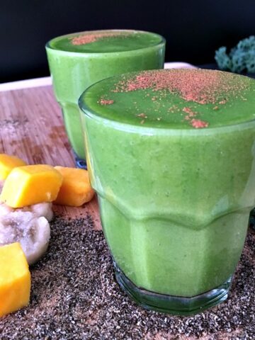 Two glasses of green smoothie sit on a bamboo board, surrounded by ingredients in the smoothies: frozen banana slices, frozen mango, chia seeds, and fresh kale leaves.