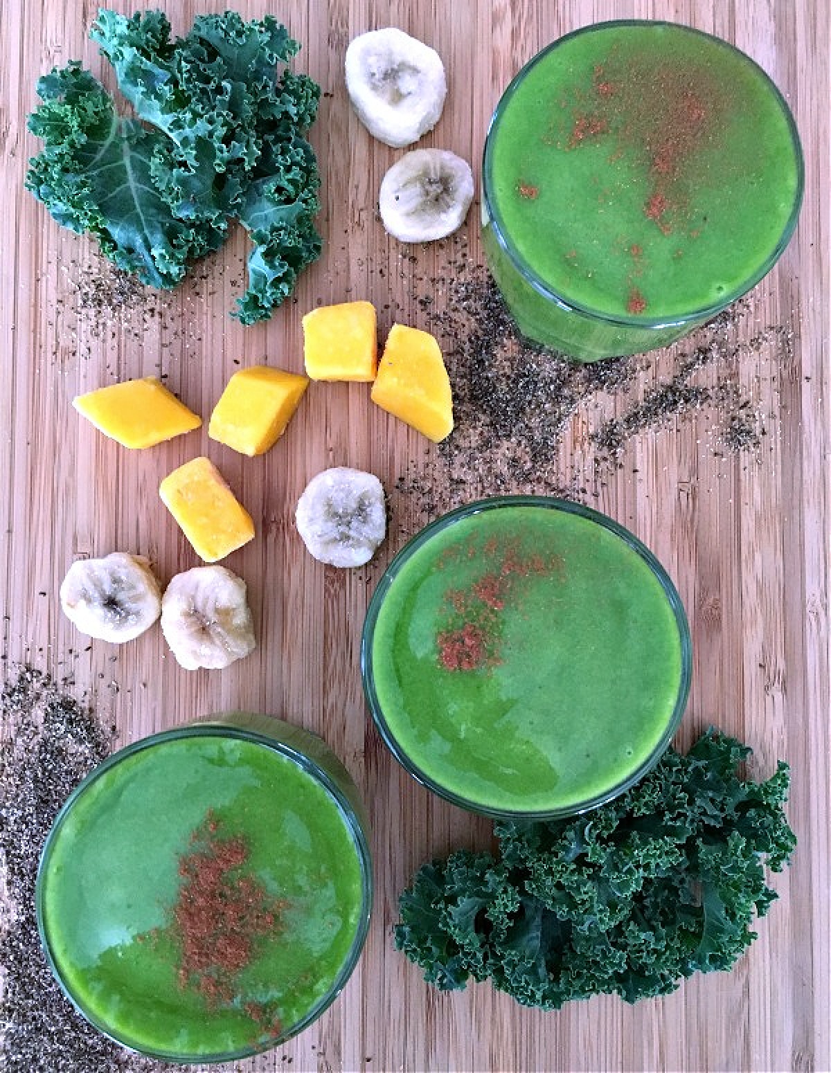 Overhead view of three glasses of green smoothie on a bamboo board, surrounded by ingredients in the smoothies: frozen banana slices, frozen mango, chia seeds, and fresh kale leaves.