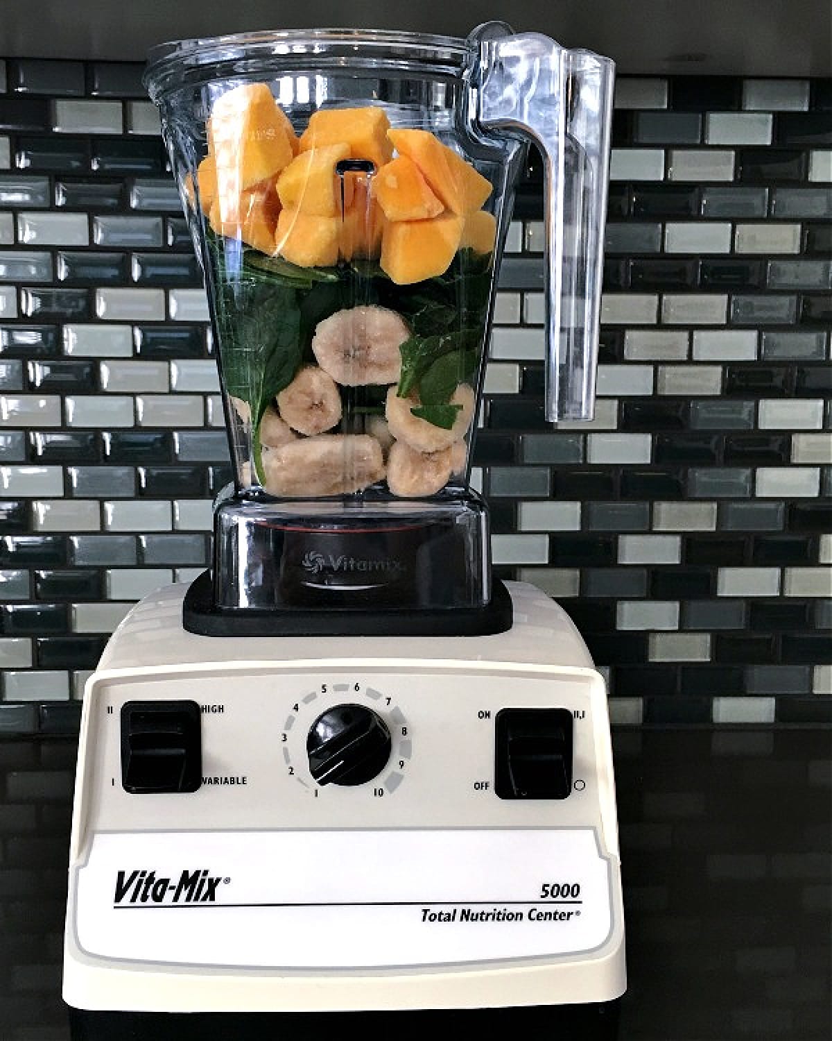 A white Vitamix blender sits on a dark grey counter against a grey glass tile backsplash. The jar of the blender is filled with smoothie ingredientsL frozen banana slices, fresh spinach, frozen mango, fresh ginger.