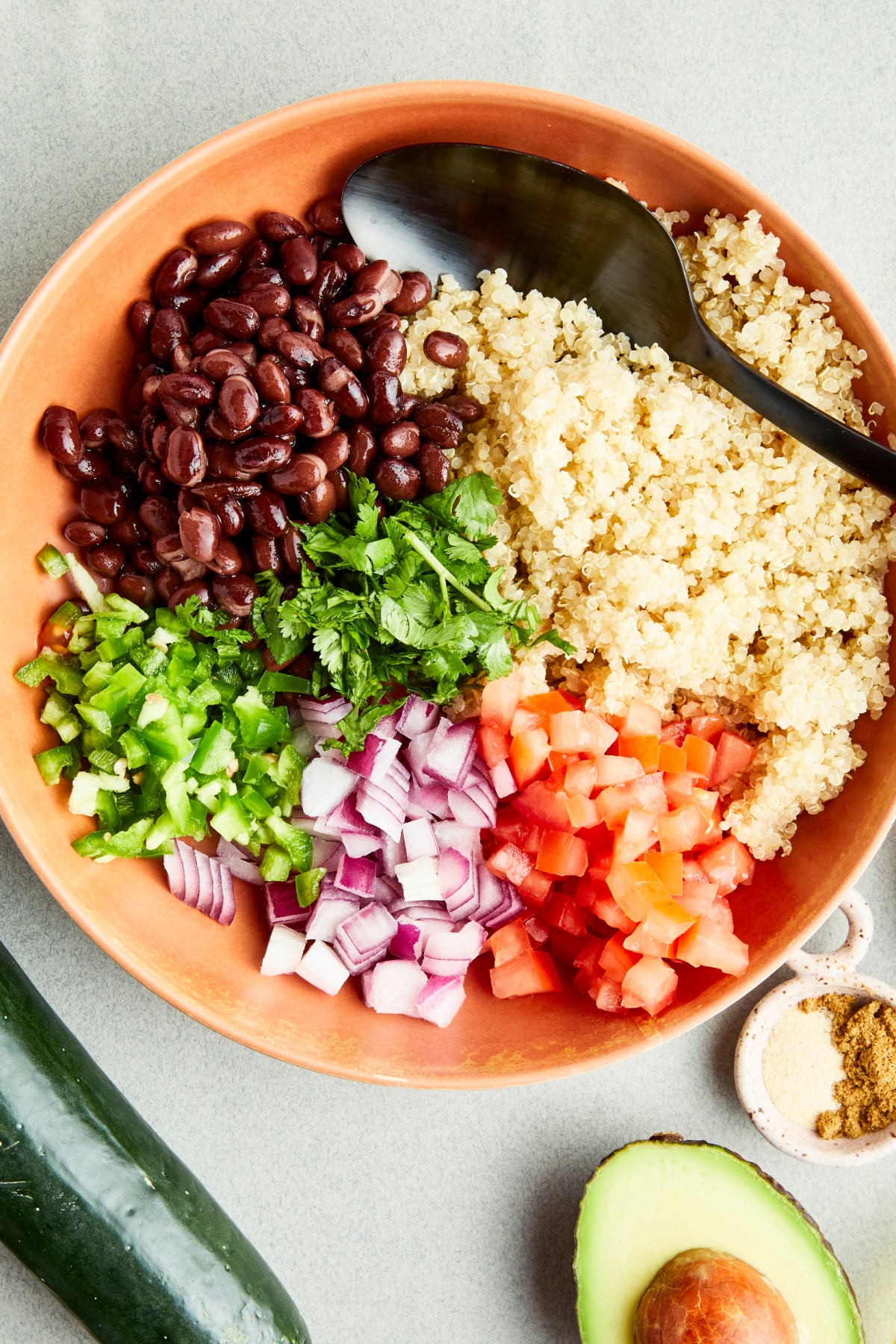 Overhead view of a terracotta colored bowl holds cooked black beans, quinoa, tomato, onion, jalapeno, and herbs before mixing. A whole cucumber, a small bowl of spices, and a half avocado sit next to the bowl.