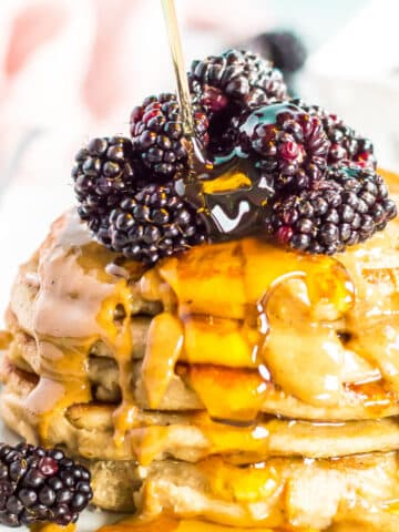 A stack of egg free applesauce pancakes on a white plate. Pancakes are topped with peanut butter, syrup, and blackberries.