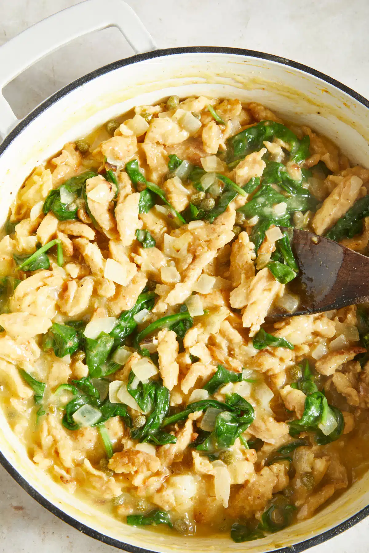 Overhead view of a white Dutch oven filled with vegan chicken piccata, which is soy curls dredged, sautéed, then cooked in a lemon caper sauce with spinach.