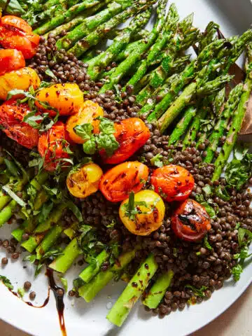 An overhead view of caprese grilled asparagus and lentils on a serving platter; asparagus is covered in lentils and grilled cherry tomatoes, fresh basil and a drizzle of balsamic vinegar.