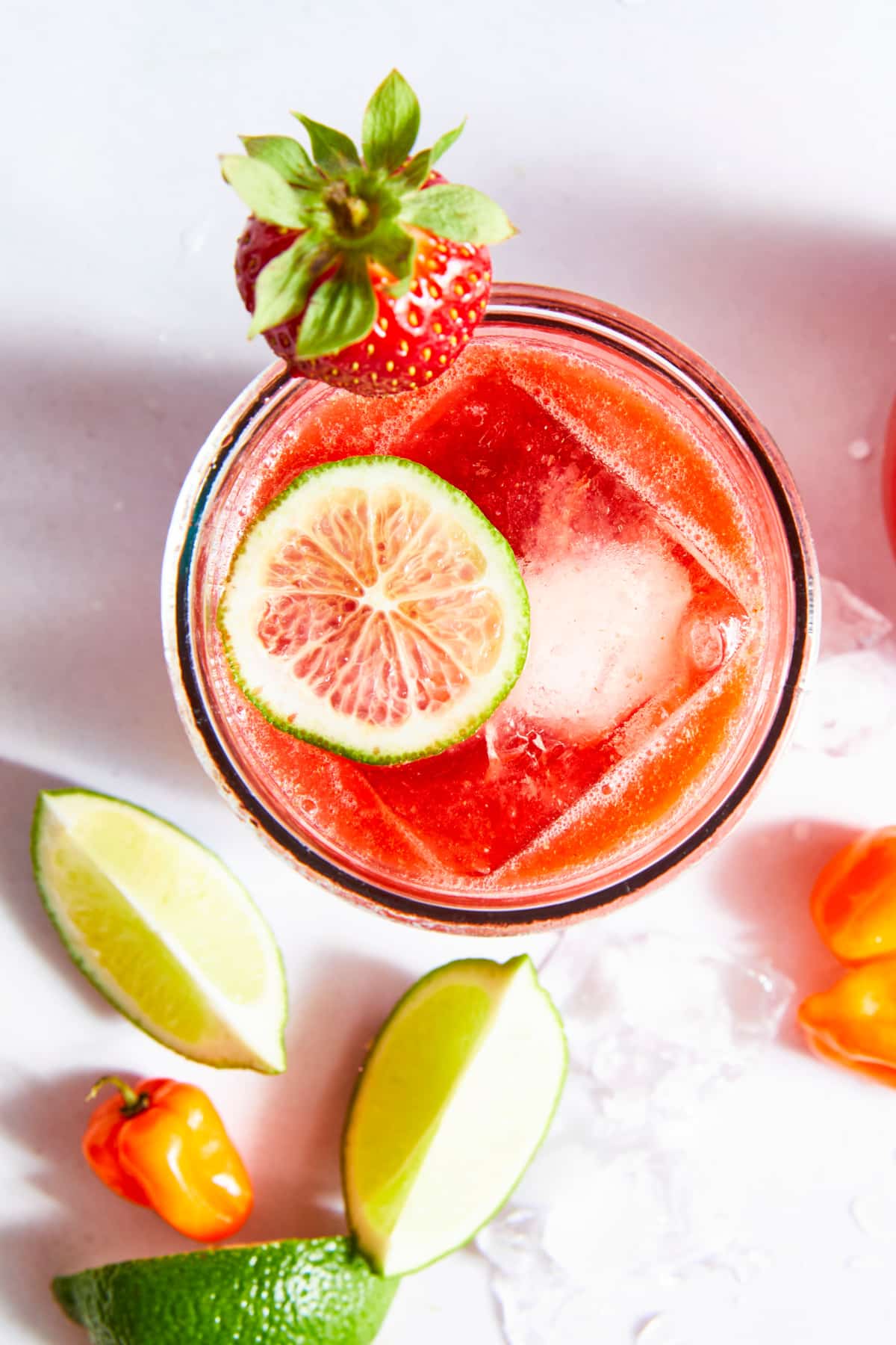 Overhead view of a spicy strawberry habanero margarita. The glass is garnished with a strawberry and surrounded by ice, habanero peppers and lime slices.