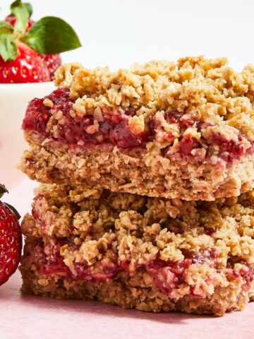Two strawberry jam bars sit stacked on a light pink surface, in front of a small white bowl of fresh strawberries.