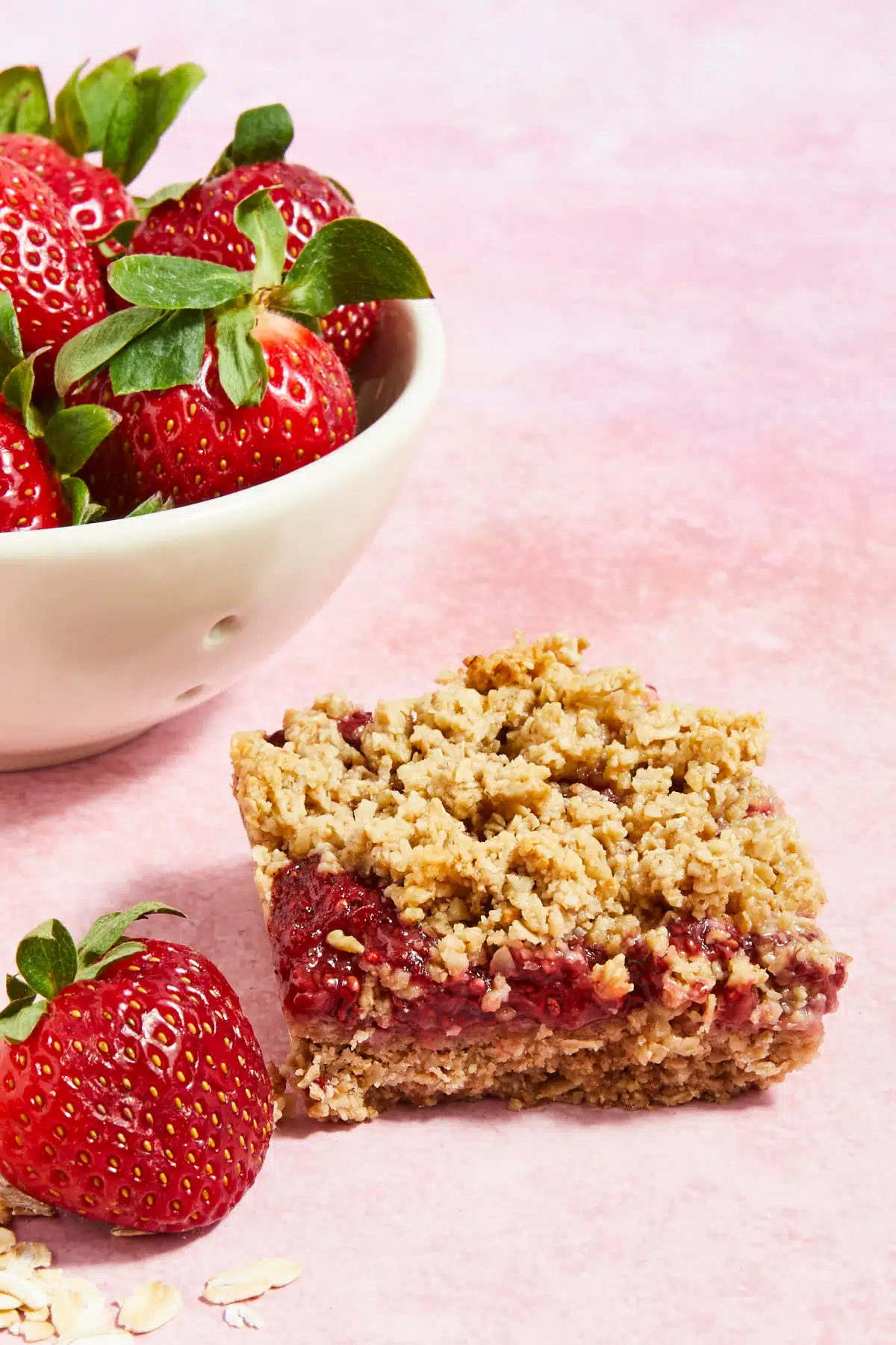 A strawberry jam crumble bar sits on a light pink surface, in front of a small white bowl of fresh strawberries.