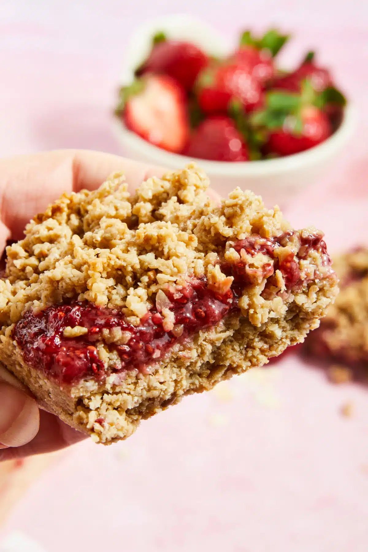 A hand holds a strawberry jam crumble bar, with a small white bowl of fresh strawberries blurred in the background.