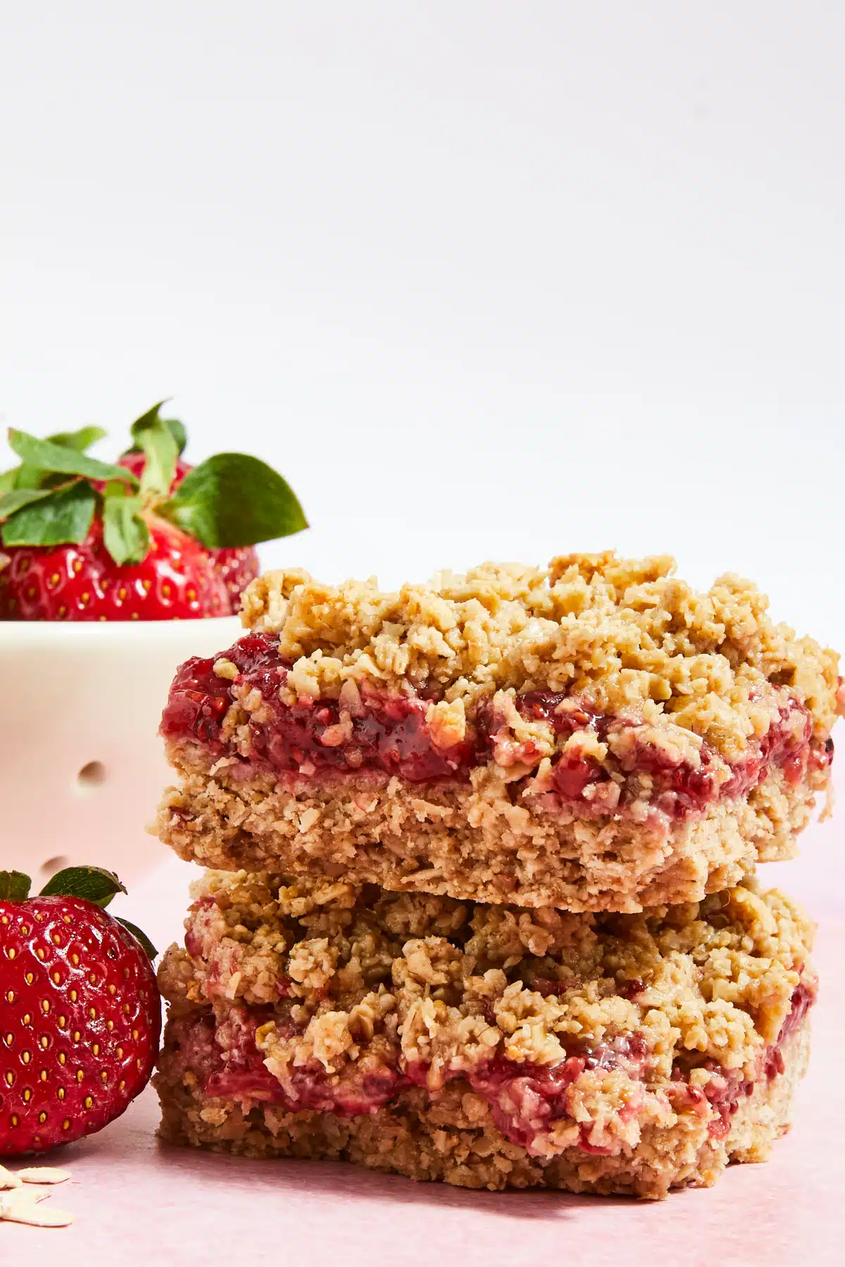 Two peanut butter jelly bars sit stacked on a light pink surface, in front of a small white bowl of fresh strawberries.