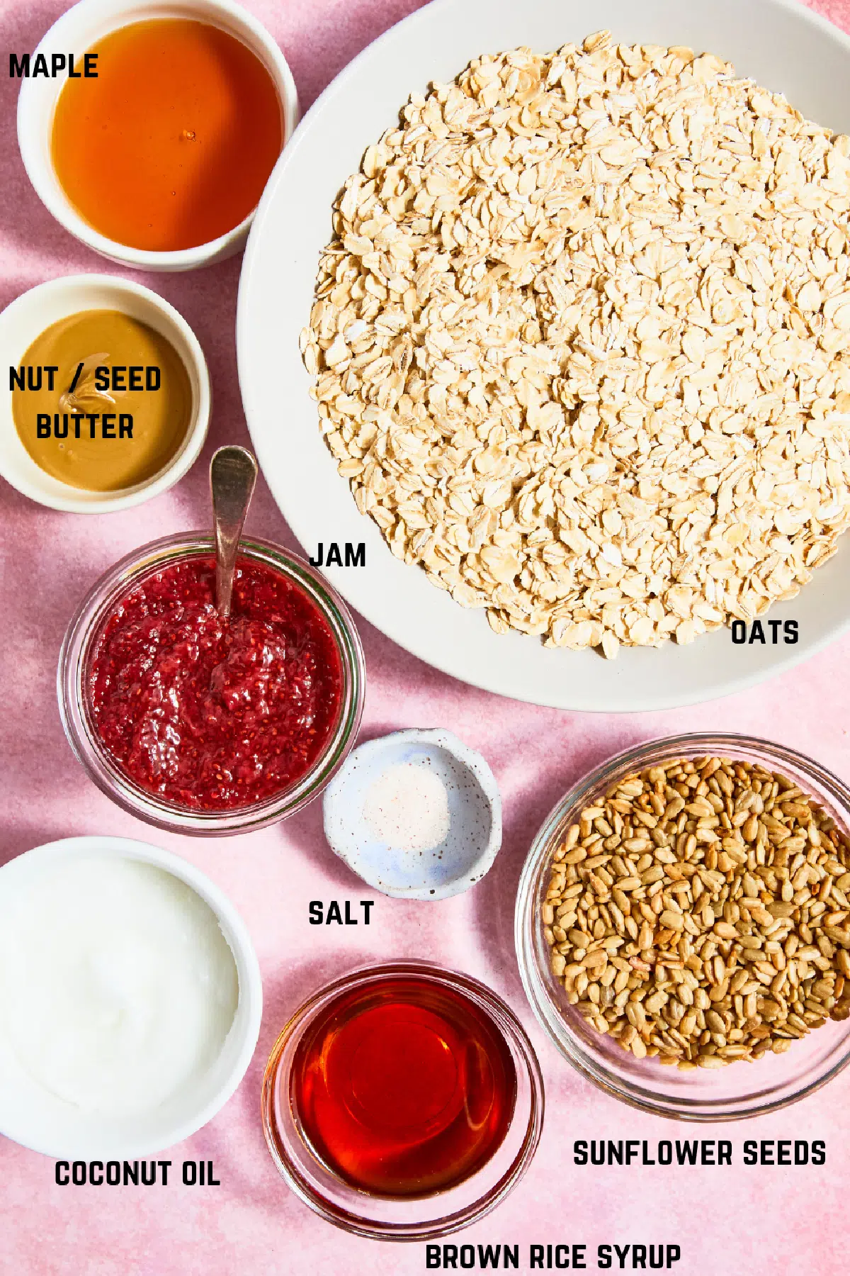 Ingredients to make strawberry oat bars: maple syrup, oats, nut butter, jam, salt, coconut oil, brown rice syrup, and sunflower seeds.