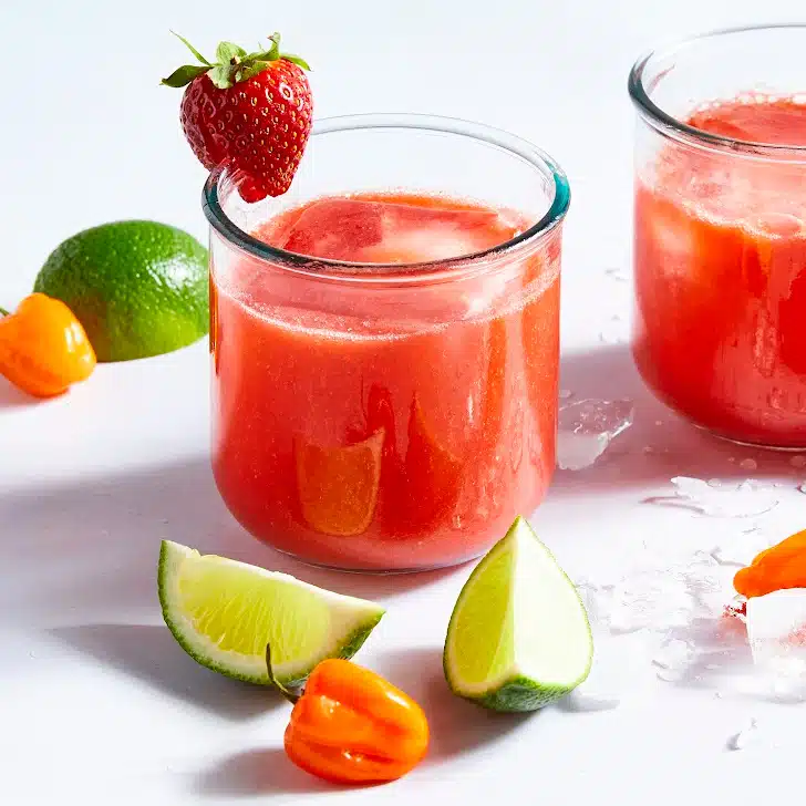 Two glasses of bright red strawberry habanero margarita. The glass is garnished with a strawberry and surrounded by habaneros and lime slices.