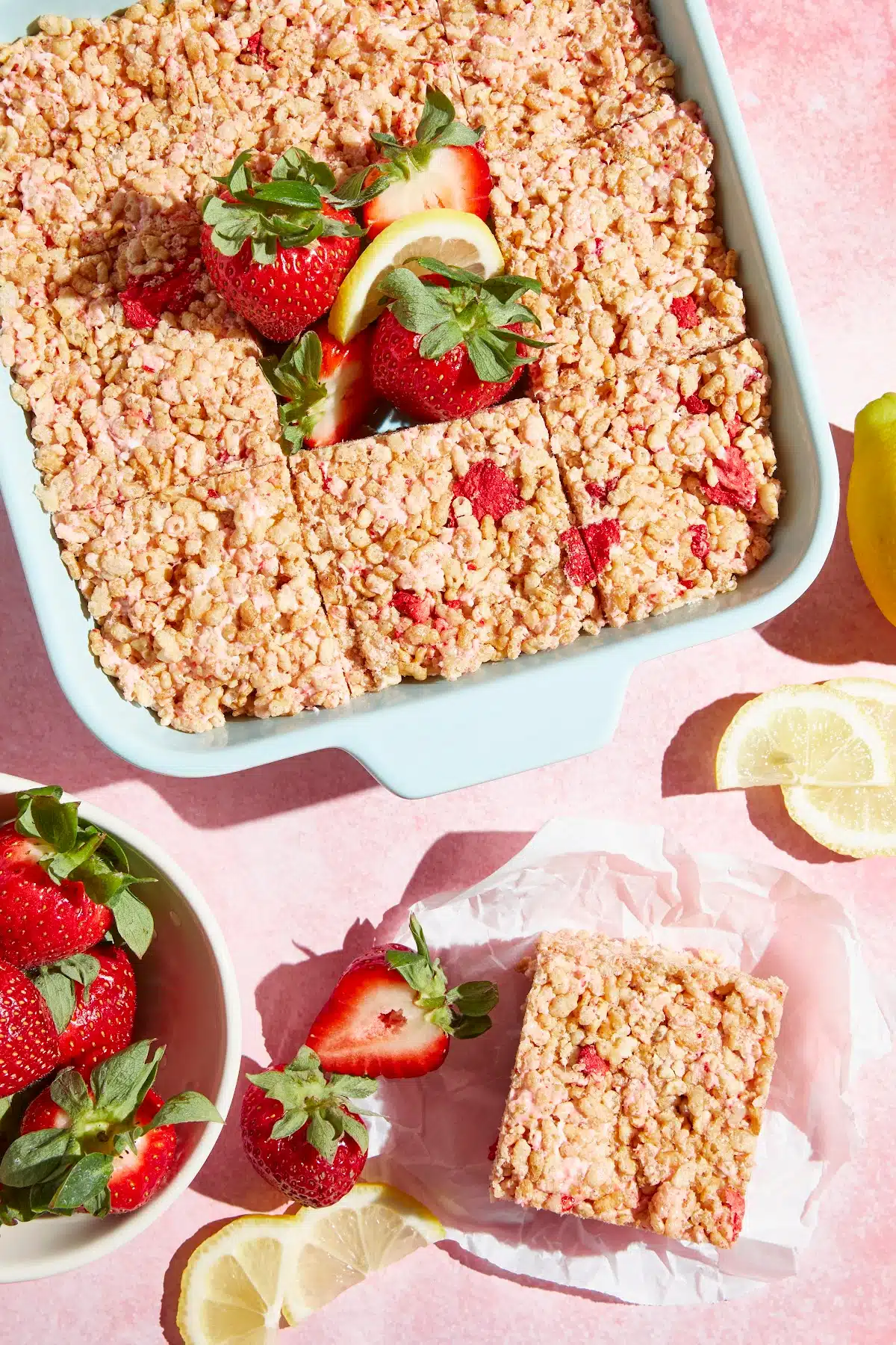 Overhead view of a pan of strawberry lemon rice crispies with one square cut out and sitting on the side. Fresh strawberries and lemon slices are scattered around and also in the space where the single square was removed from the pan.