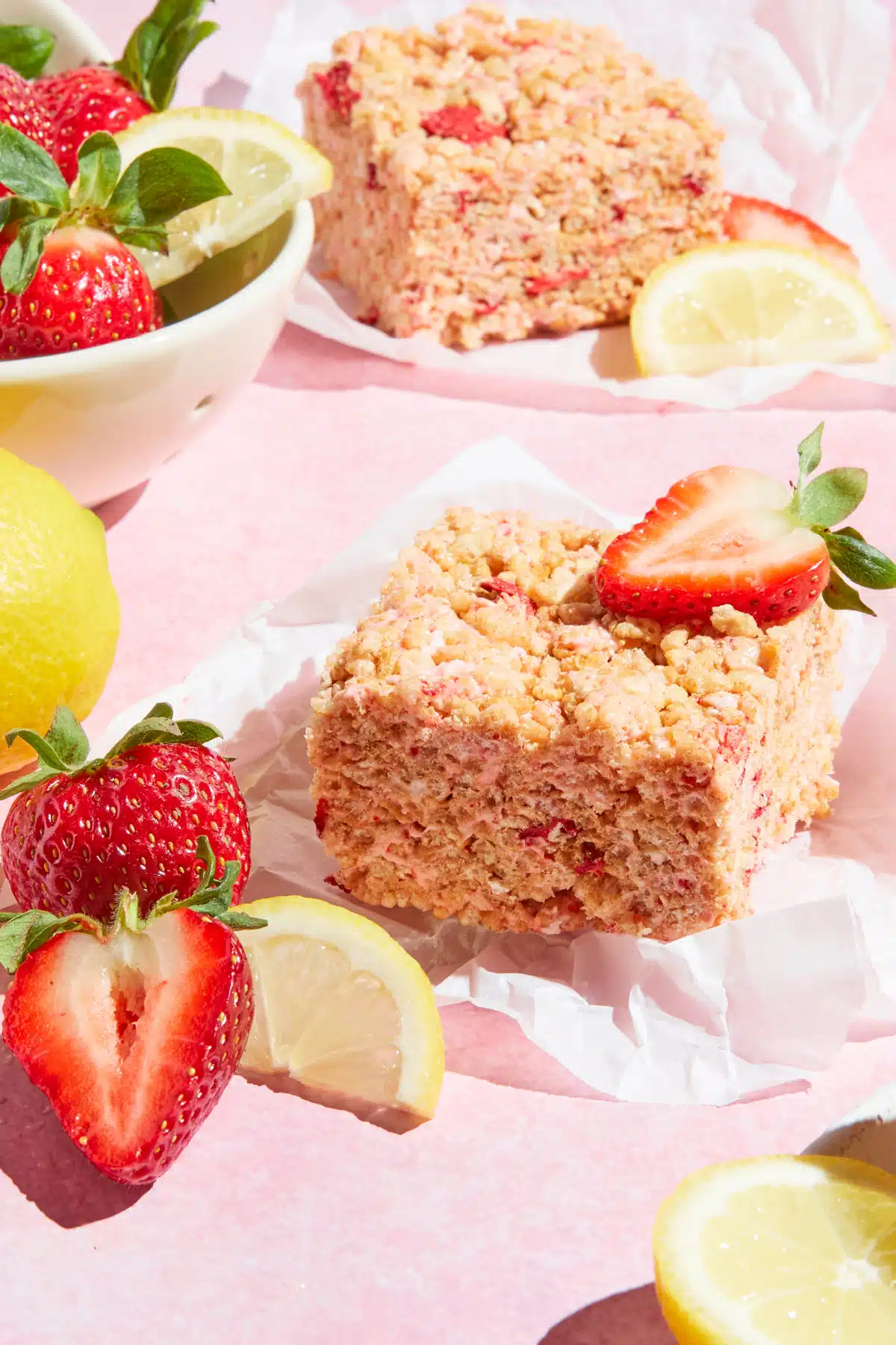 Two strawberry lemon rice crispies squares on small pieces of parchment on a pink surface. A bowl of fresh strawberries and lemons sit on the side.