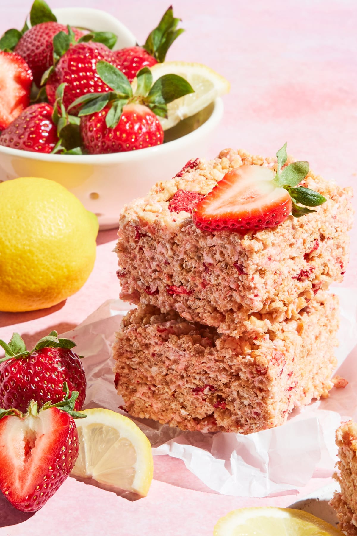 Two strawberry lemon rice crispy treat squares stacked together on a small square of parchment. A bowl of fresh strawberries and lemons sit on the side.