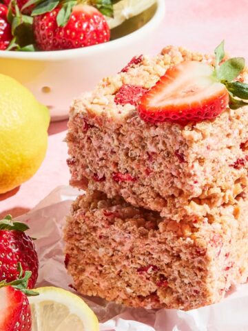 Two strawberry lemon rice crispy treat squares stacked together on a small square of parchment. A bowl of fresh strawberries and lemons sit on the side.