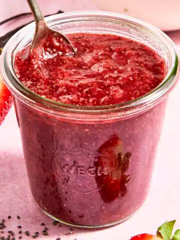 A glass canning jar of strawberry vanilla chia jam with a spoon in it sits on a light pink surface. Fresh strawberries and lemon slices sit in a small white bowl next to the jam, and strawberry slices, vanilla bean, and chia seeds are sitting around the jam jar.