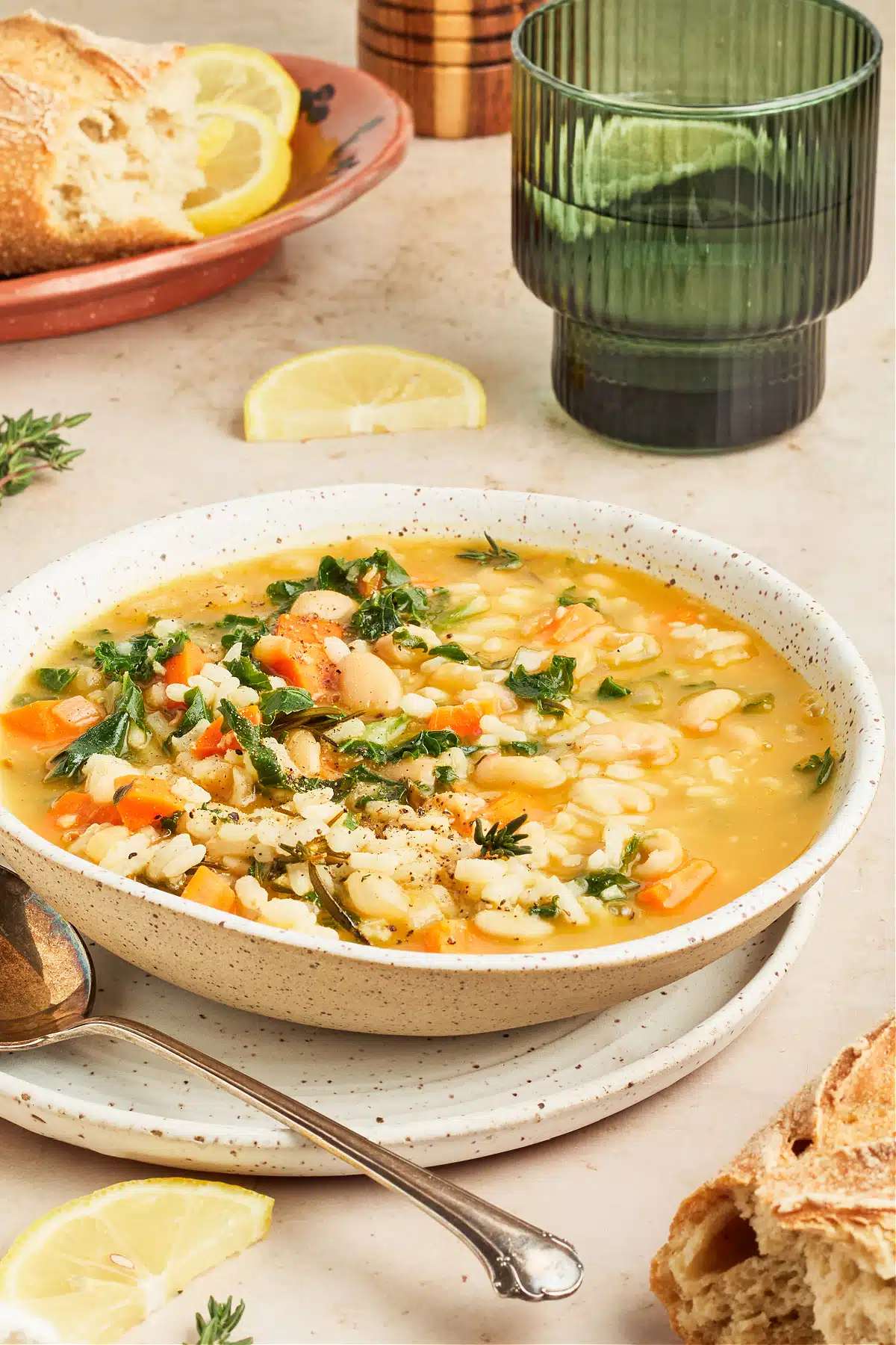 A bowl of lemon bean soup with rice and kale sits on a white surface with a green glass of wine and a plate of crusty bread. A spoon is in the bowl and there is a lemon wedge on the side.