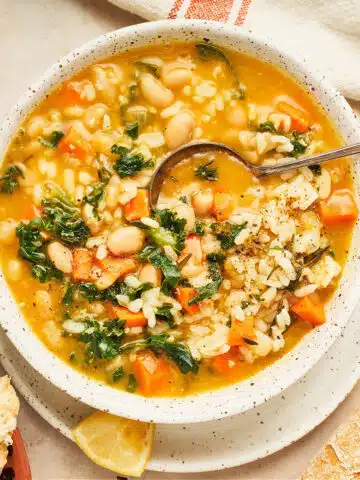 Overhead view of a bowl of lemon soup with rice, kale, and beans. A spoon is in the bowl and there is a lemon wedge and crusty bread on the side.