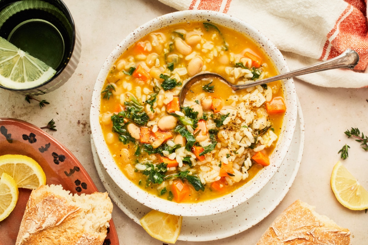 A bowl of vegetable soup filled with white beans, rice, kale, carrots, and broth. A spoon rests in the bowl. The setting includes a piece of rustic bread, lemon wedges, and a green glass of water with a lime wedge. A cloth napkin lies beside the bowl.
