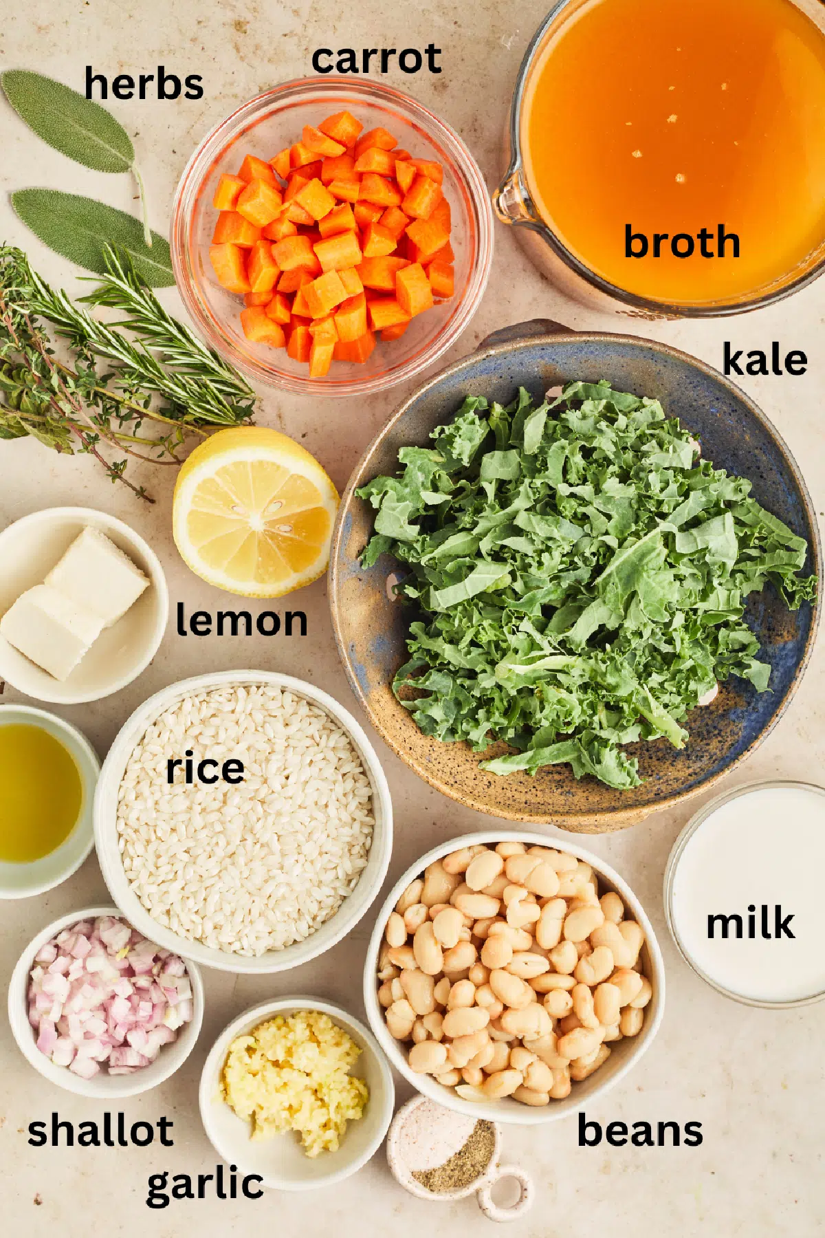 Overhead view of ingredients to make lemon kale white bean soup: fresh herbs, chopped carrots, broth, chopped kale, lemons, rice, minced shallot, minced garlic, white beans, and dairy free milk.