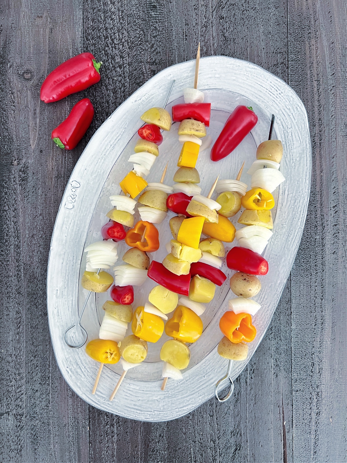 Overhead view of a large platter holding potato veggie skewers before grilling. Skewers hold mini gold potatoes, sliced onion, and mini sweet peppers.