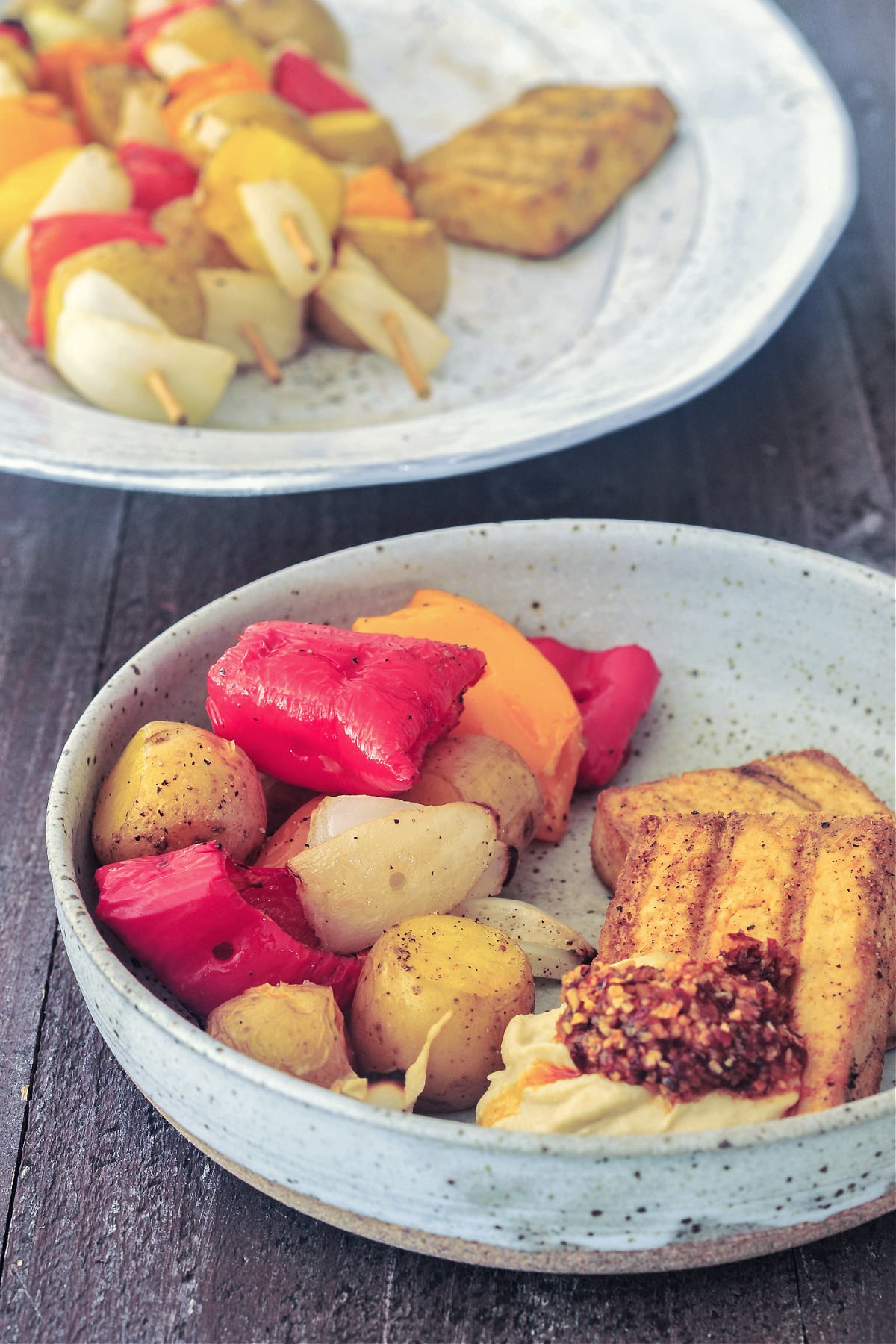 A rustic light grey bowl of grilled veggies (peppers, onions, and mini potatoes) and grilled tofu slabs garnished with hummus and chili crisp sit on a wood table. A platter of grilled veggie skewers sit in background.