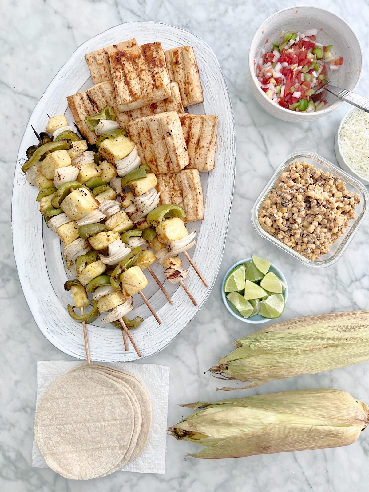 Overhead view of a white marble tabletop holding plates of grilled food: a platter of grilled veggie skewers filled with green peppers, onions, and pineapple and slabs of grilled tofu, a bowl of fresh homemade salsa, two grilled cobs of corn, a small dish of lime wedges, and a pile of corn tortillas.