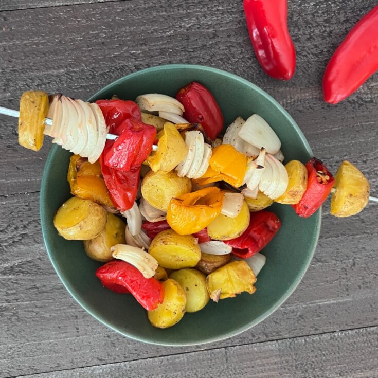 Overhead view of grilled veggies (peppers, onions, and mini potatoes) in a green bowl on a wood table. two raw mini red sweet peppers sit next to the bowl. One skewer fresh from the grill sits over the bowl, filled with these grilled veggies.