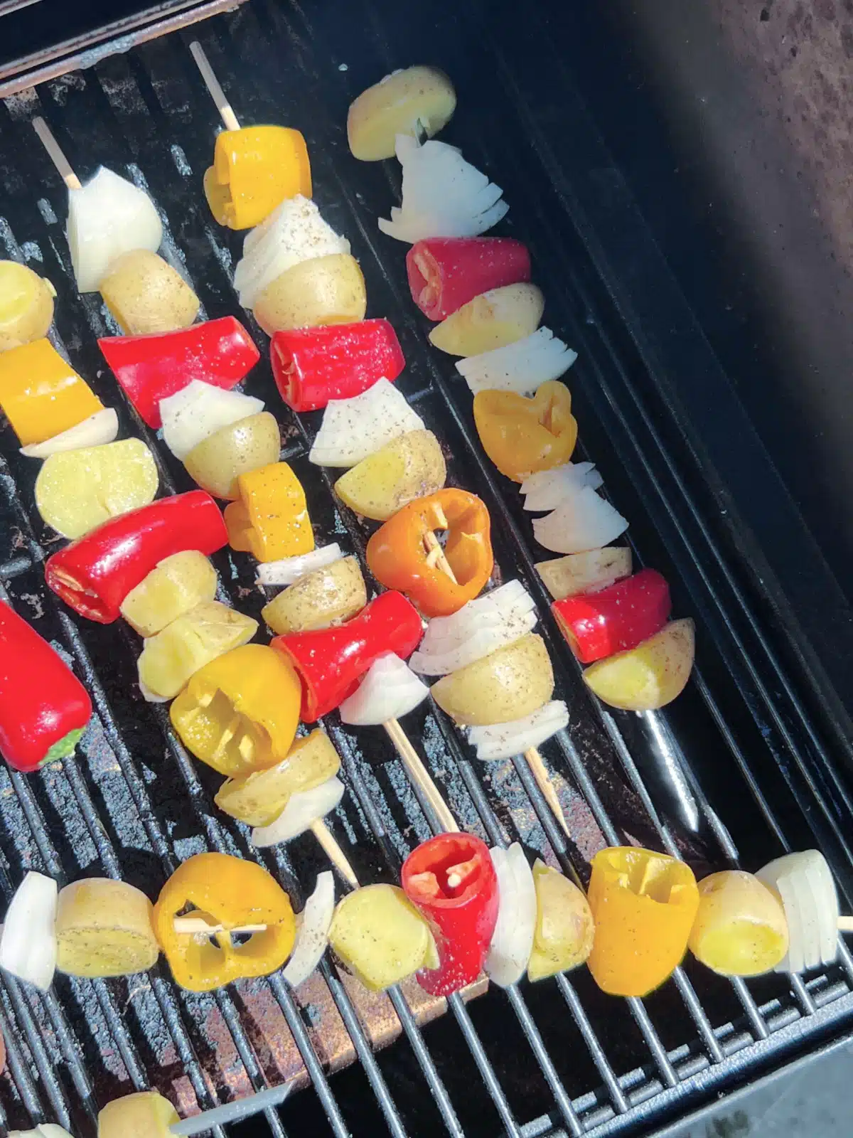 Potato veggie skewers sit on a grill, cooking in the sunshine. The skewers have mini gold potatoes, onion quarters, and mini sweet peppers.