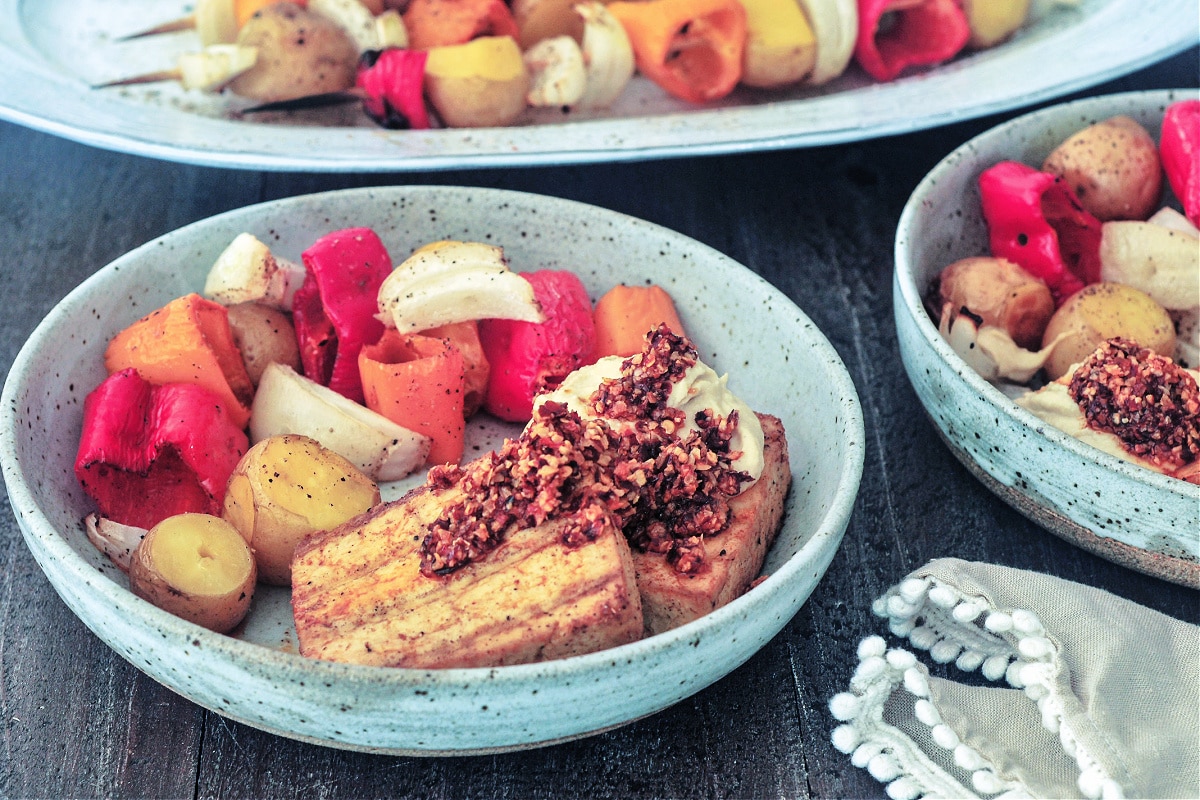 Two rustic light grey bowls of grilled veggies (peppers, onions, and mini potatoes) and grilled tofu slabs garnished with hummus and chili crisp sit on a wood table. A platter of grilled veggie skewers sit in background.