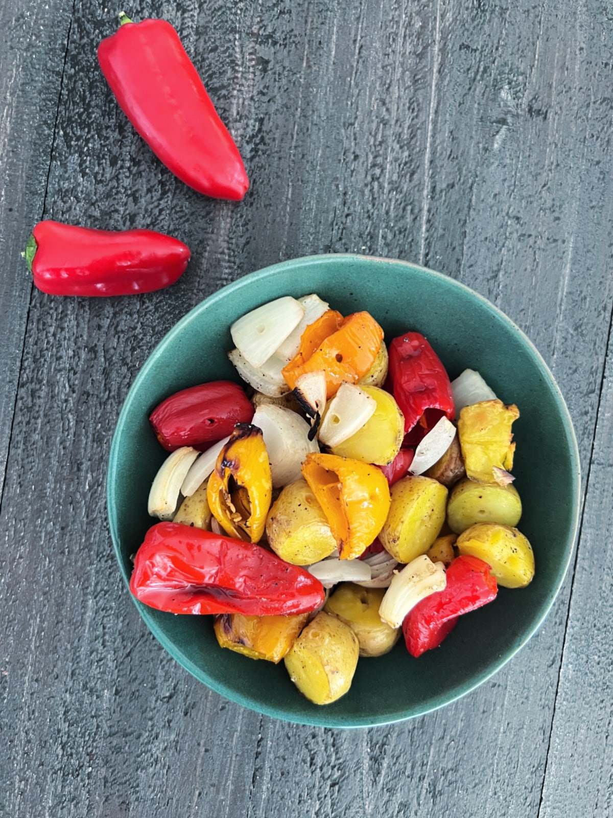 Overhead view of grilled veggies (peppers, onions, and mini potatoes) in a green bowl on a wood table. two raw mini red sweet peppers sit next to the bowl.