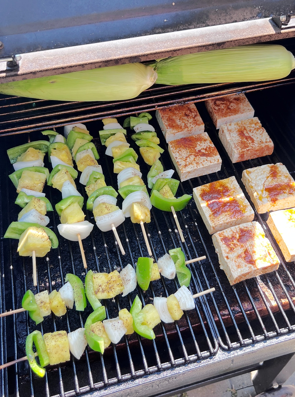 Veggie skewers sit on a grill, cooking in the sunshine. The skewers have pineapple cubes, onion quarters, and mini sweet peppers. The grill also holds tofu slabs marinated in a smoked paprika olive oil mixture, and two cobs of corn in their husks.