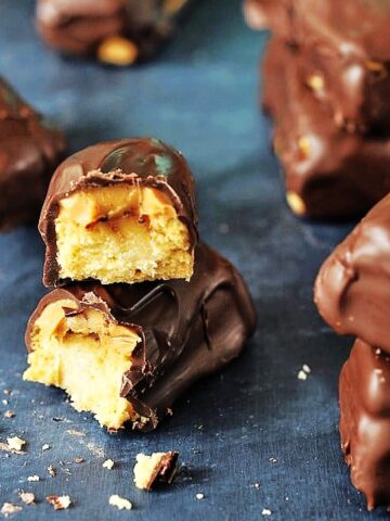 Close up image of a homemade peanut butter Twix bar broken in half to see the peanut butter and cookie inside. Several chocolate covered bars sit in background.