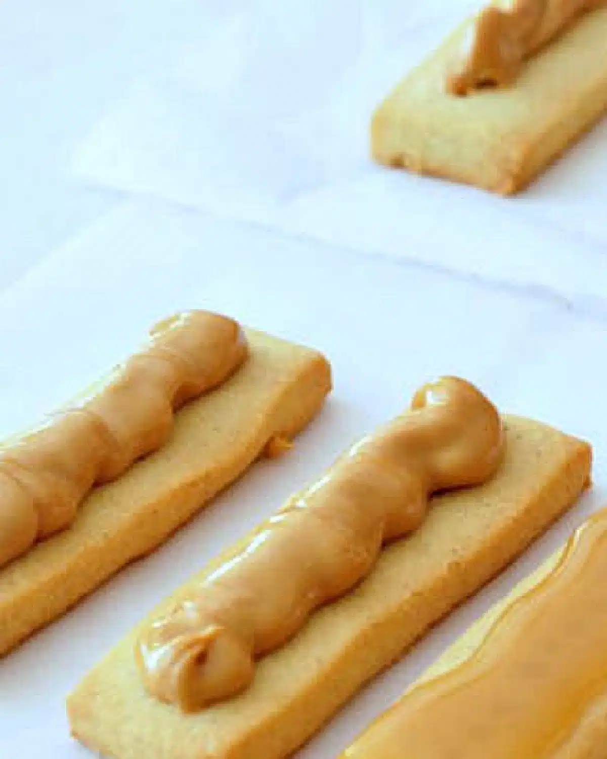 Several long shortbread cookies spread with peanut butter, in an image showing a step in how to make homemade peanut butter Twix bars.