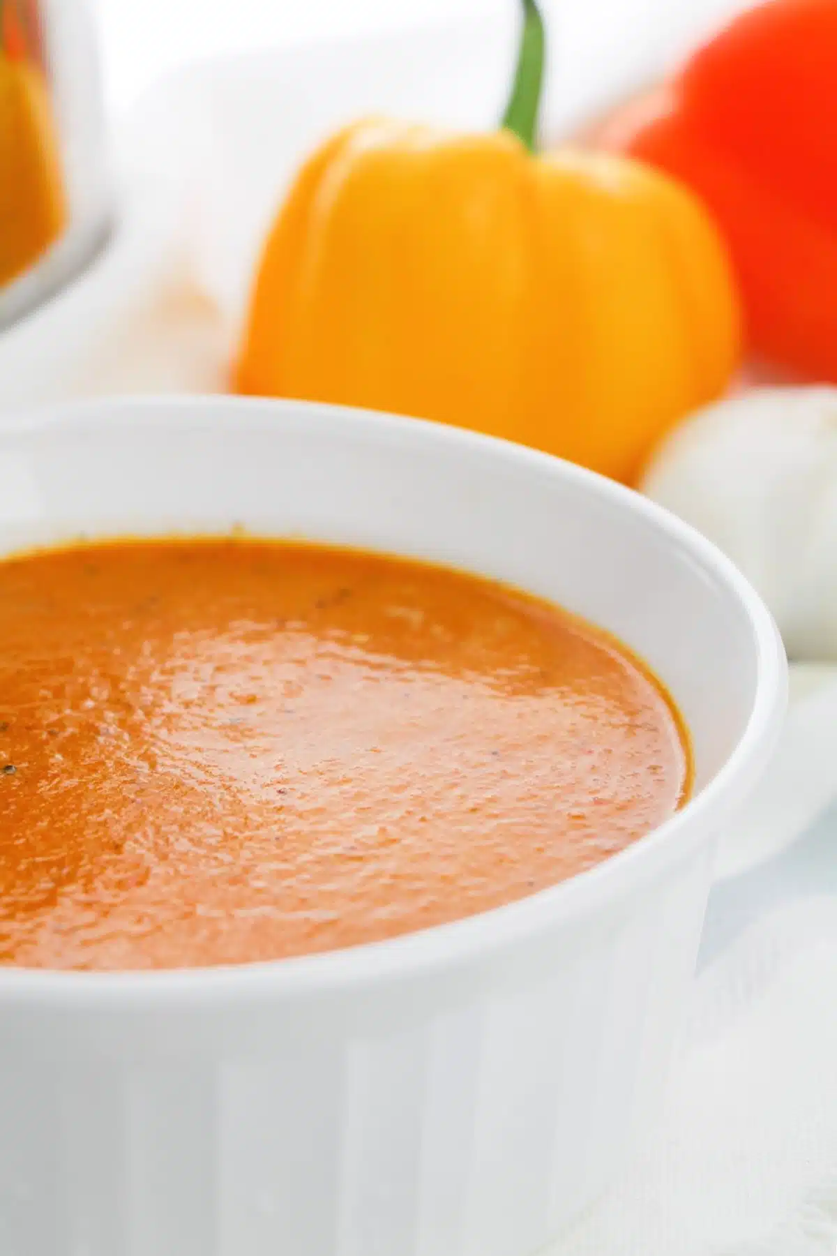 Roasted pepper bisque is served in a bright white bowl. Whole bell peppers and a garlic bulb are in the background.